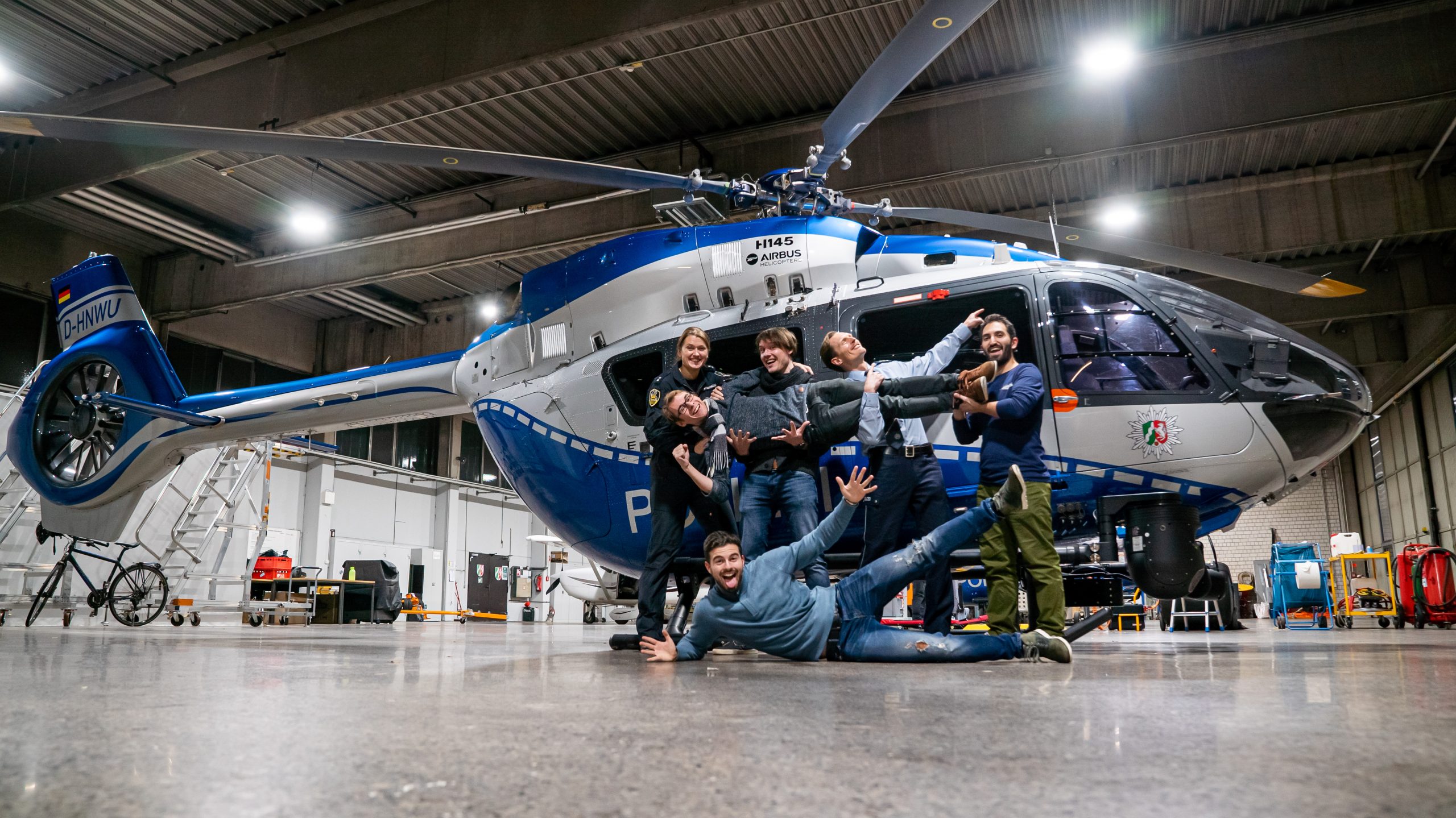 Sechs Personen posieren vor einem Helikopter der Polizei. Der blau-silberne Helikopter ist in voller Länge abgebildet.