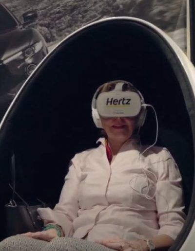 Eine Frau sitzt in einem kugelförmigen Stuhl und hat eine VR Brille mit der Aufschrift Hertz auf. Im Hintergrund ist eine Wand mit einem großen Foto eines Maserati.