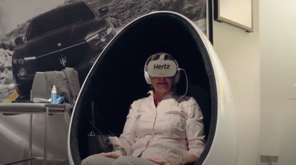Eine Frau sitzt in einem kugelförmigen Stuhl und hat eine VR Brille mit der Aufschrift Hertz auf. Im Hintergrund ist eine Wand mit einem großen Foto eines Maserati.