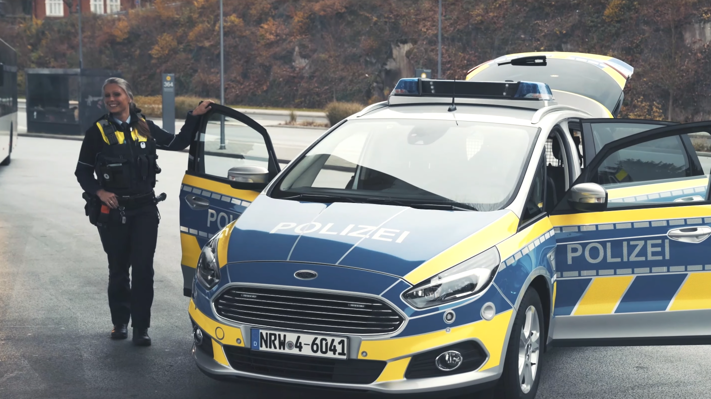 Blau-gelbes Polizeiauto steht mit geöffneten Türen auf der Straße. Links von dem Auto steht eine Polizistin mit ihrer Hand auf de Kante der Autotür.