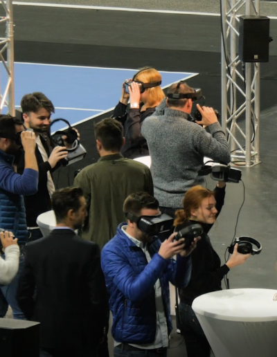 Ein Gruppe von Personen sind um Stehtische verteilt und haben VR Brillen und Headsets in der Hand oder auf dem Kopf.